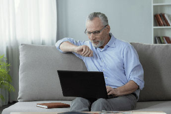 A pensioner sitting on a sofa and using one of Telstras NBN plans for seniors to connect to the internet using his laptop.
